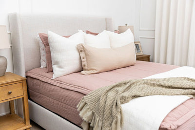 A brightly lit bedroom with a bed made with woven pink zipper bedding and an assortment of white and tan pillows and matching blankets.