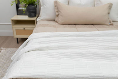 A close up of a bed made with tan zipper bedding and neutral pillows and a white piano key quilted design blanket at the end of the bed.