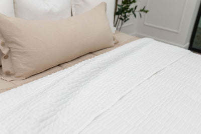 A close up of a bed made with tan zipper bedding and neutral pillows and a white piano key quilted design blanket folded over the reveal the soft white minky side. 