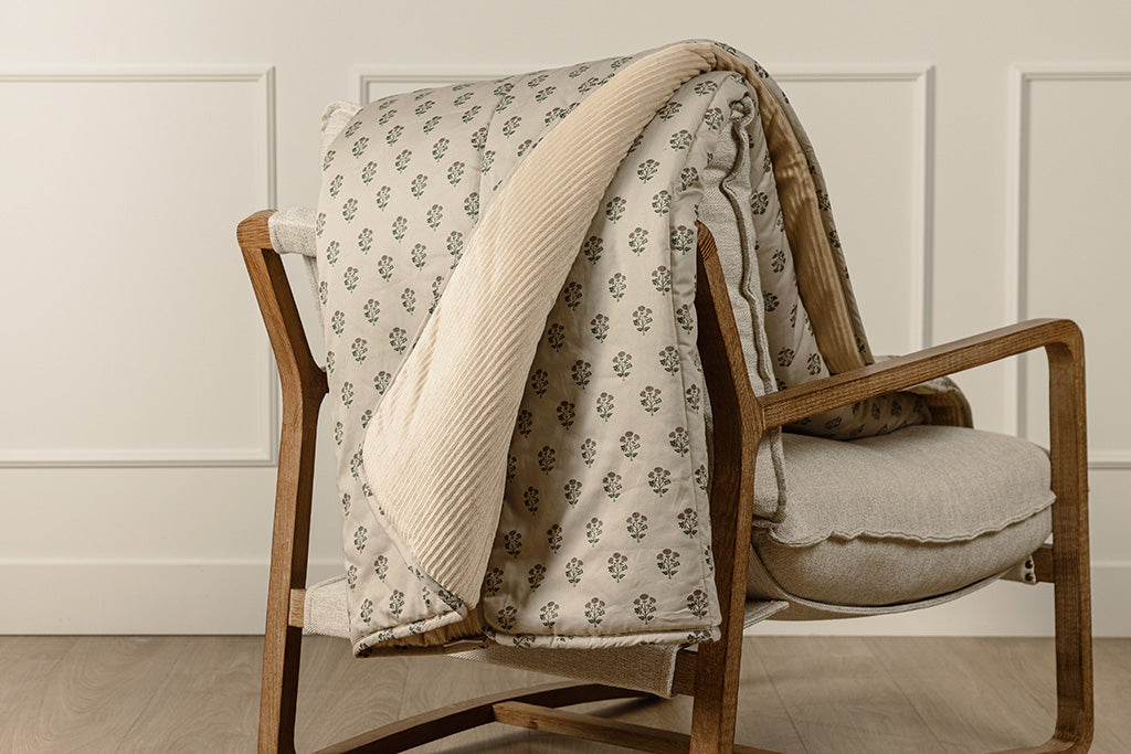 A tan, pink, and green blockprint floral  with cream corduroy minky blanket on a wood and oat colored chair.