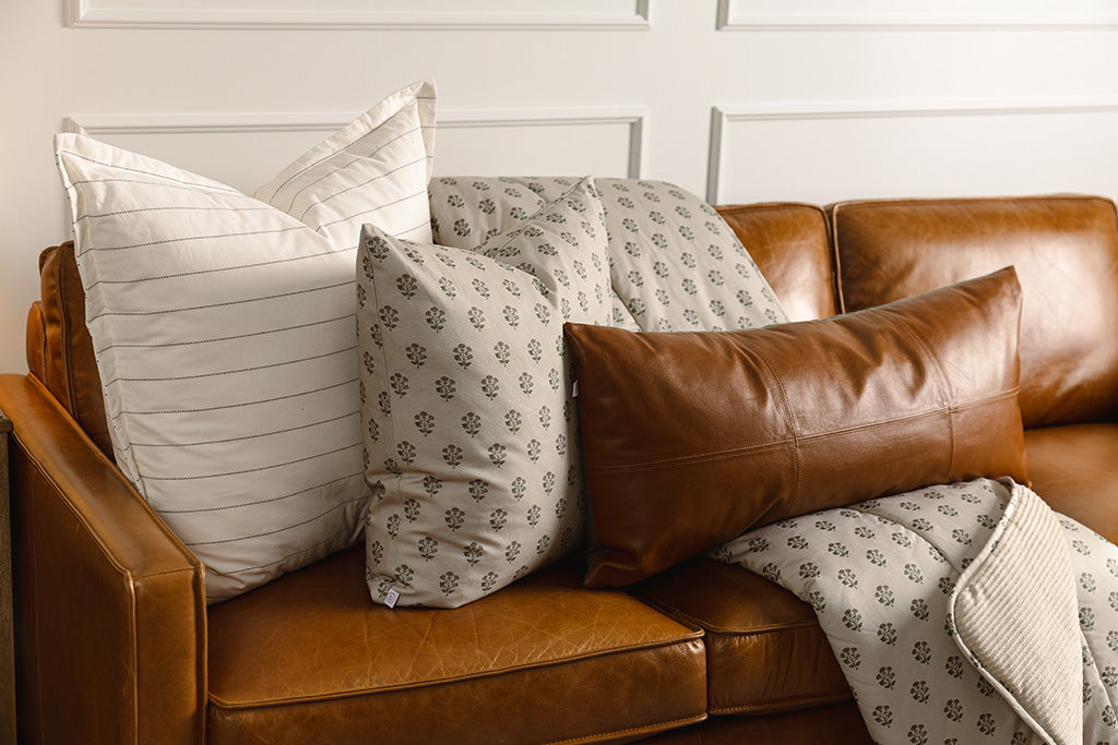 A tan, pink, and green blockprint floral pillow, white and black striped pillow, leather lumbar, and a tan, pink, and green blockprint floral blanket on a leather couch.