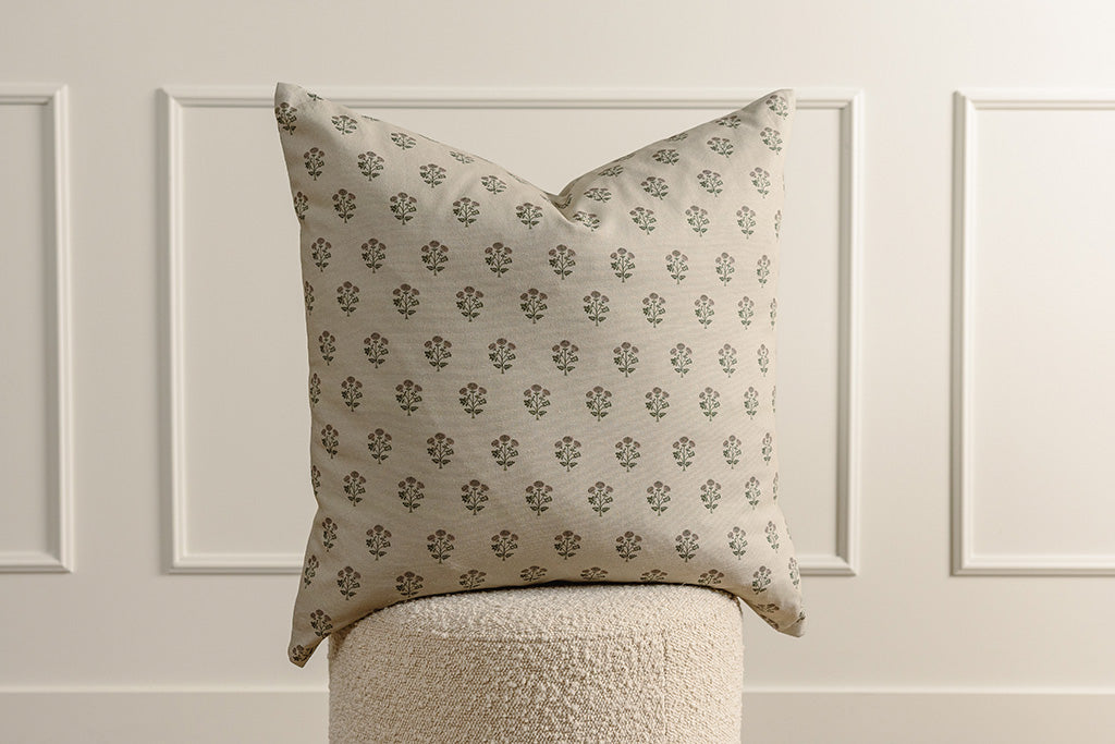 A tan, pink, and green blockprint floral pillow on a sherpa stool in front of a wainscoting background.