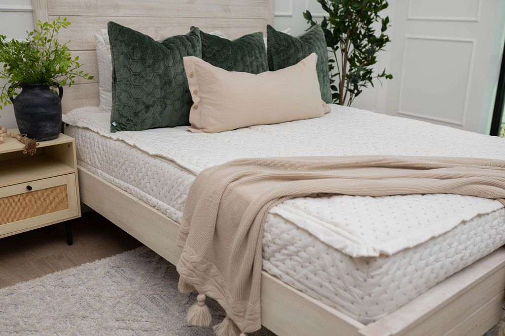 A close up of a bed made with cream embroidered zipper bedding and green velvet pillows with a tan lumbar and woven throw blanket.