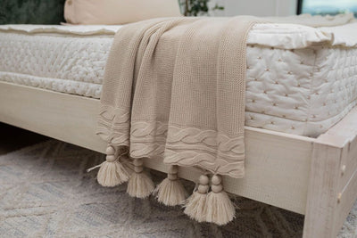A close up side view of a bed made with cream embroidered zipper bedding and a woven tan blanket with tassels at the end draped over the edge of the bed.