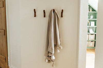 A wall with four hooks on it, one of the hooks holding a tan throw blanket with large tassels on the end.
