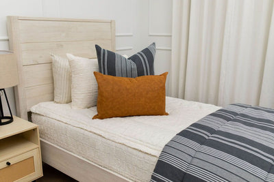 A twin bed made with cream textured zipper bedding and neutral gray and leather pillows as well as a matching striped blanket.