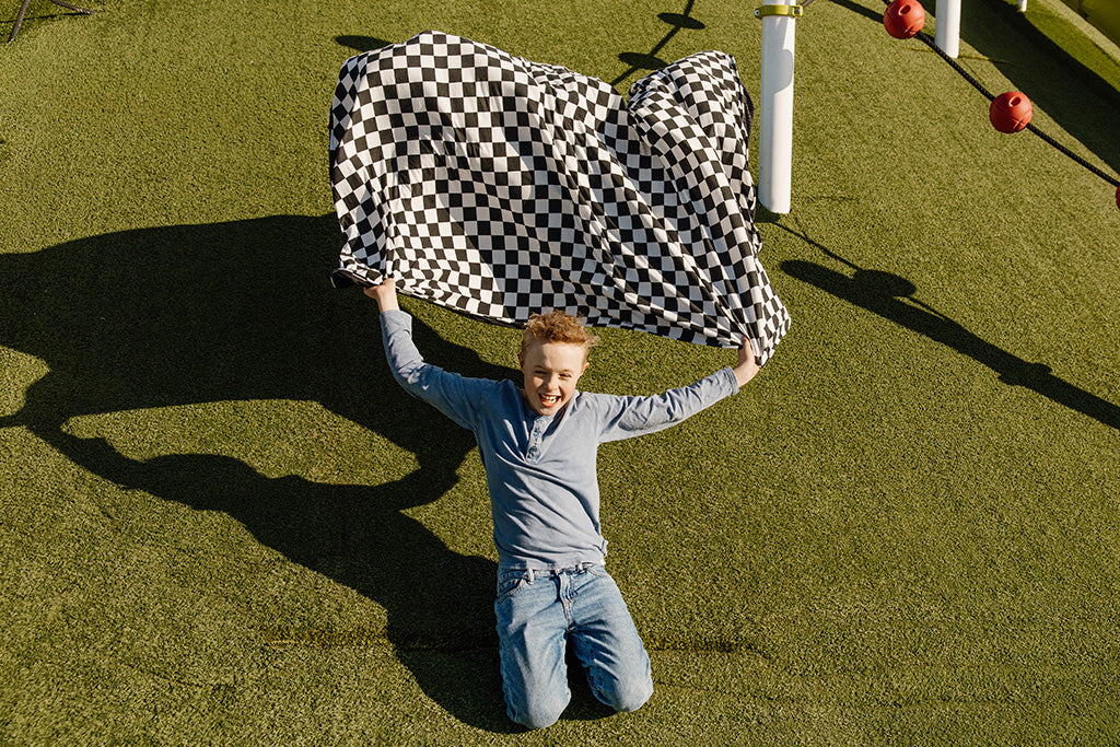 A young boy sliding down a grass hill holding a black and white checkered blanket. 