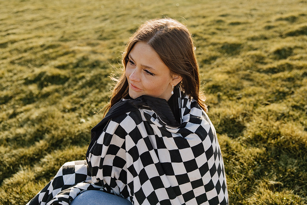 A woman wrapped in a black and white checkered blanket, sitting on a green grass field. 