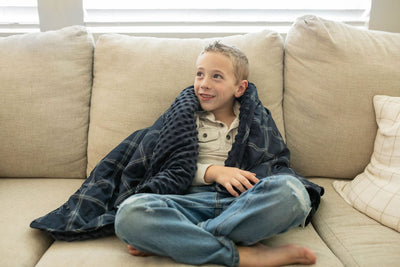 A boy smiling and sitting on couch with a navy blue plaid children's blanket wrapped around him.