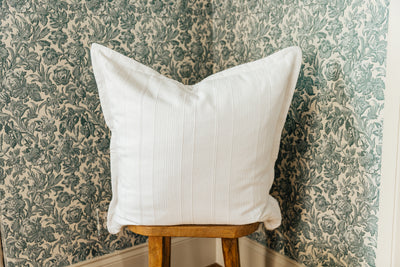 A big white textured pillow arranged on a wood stool in front of floral green wallpaper.