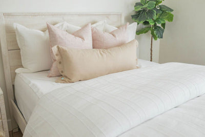 A bed made with white zipper sheets and a textured white zipper duvet folded half way down the bed and adorned with neutral and light pink pillows.