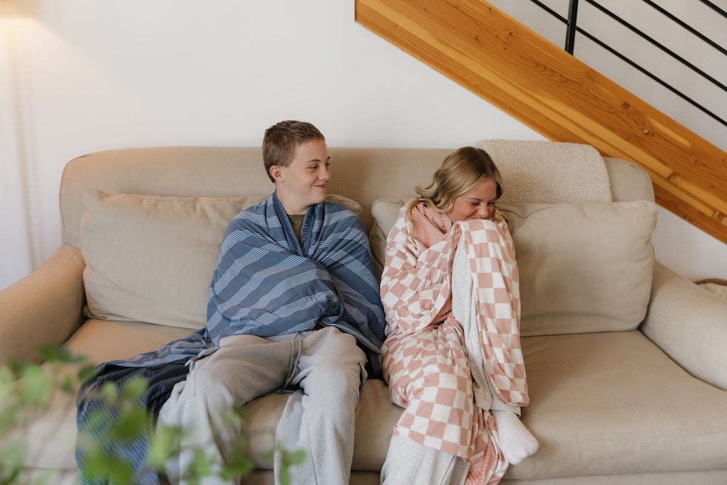 Siblings sitting on a couch cuddled in a blue and pink travel blanket. 
