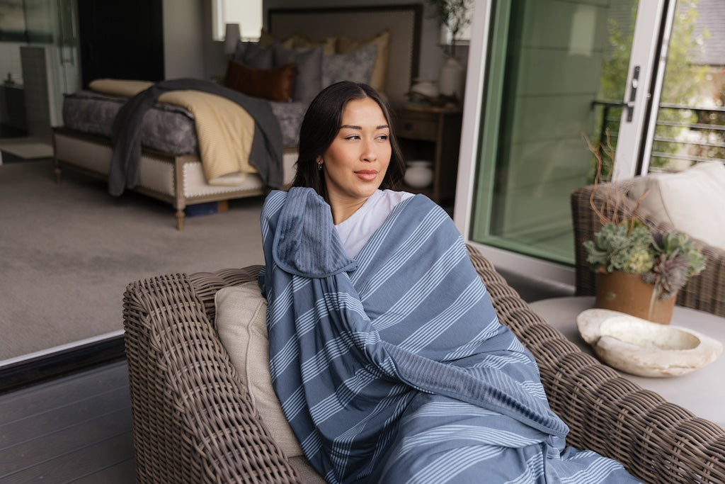 A woman sitting on a chair cuddled up in a blue stripe  travel blanket. 