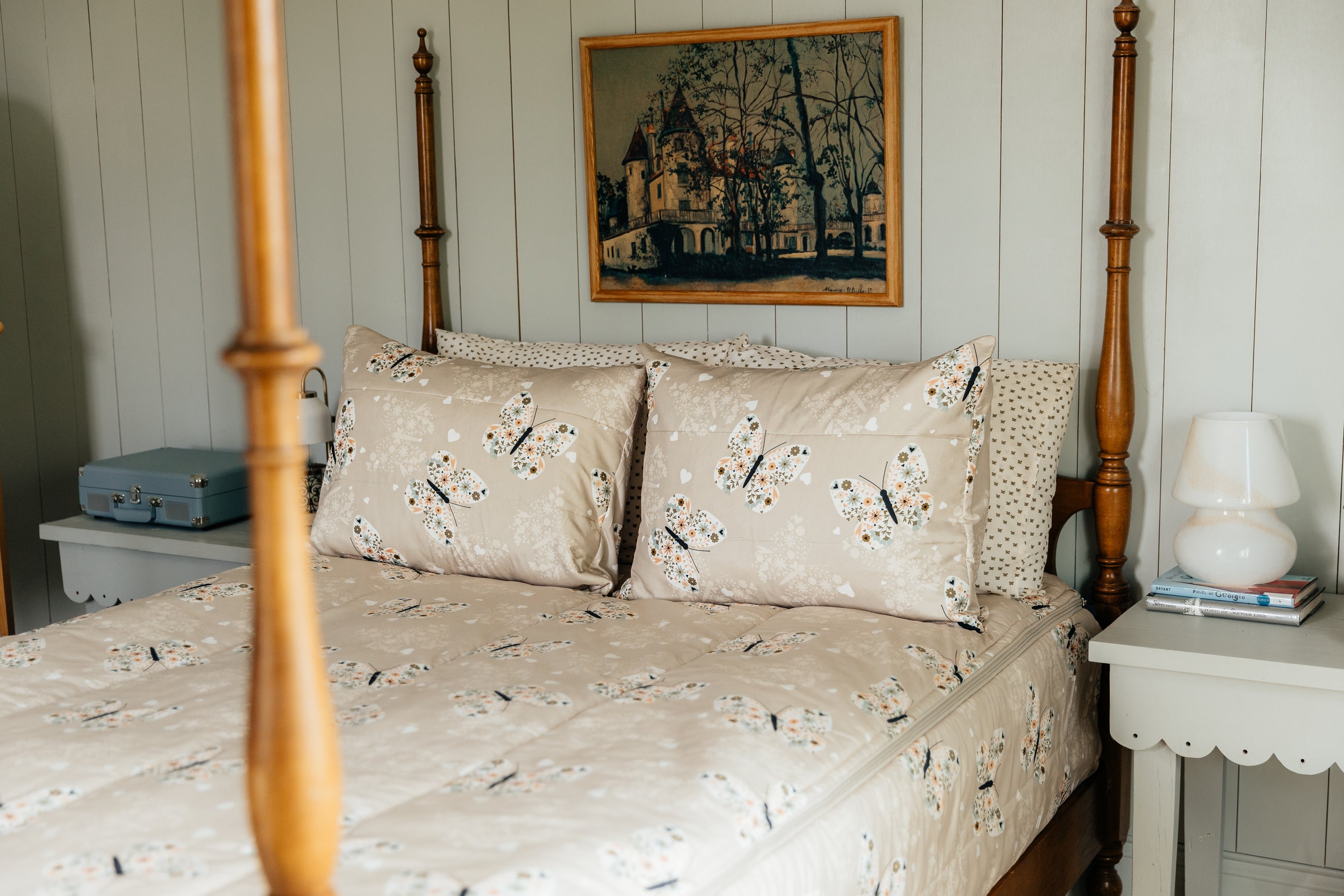 A full size four poster bed made with butterfly zipper bedding, white nightstands on either side of the bed, and a picture of a victorian house hanging over the bed.