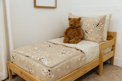 A wooden toddler bed made with butterfly printed zipper bedding and a stuffed bear leaning against the pillows.