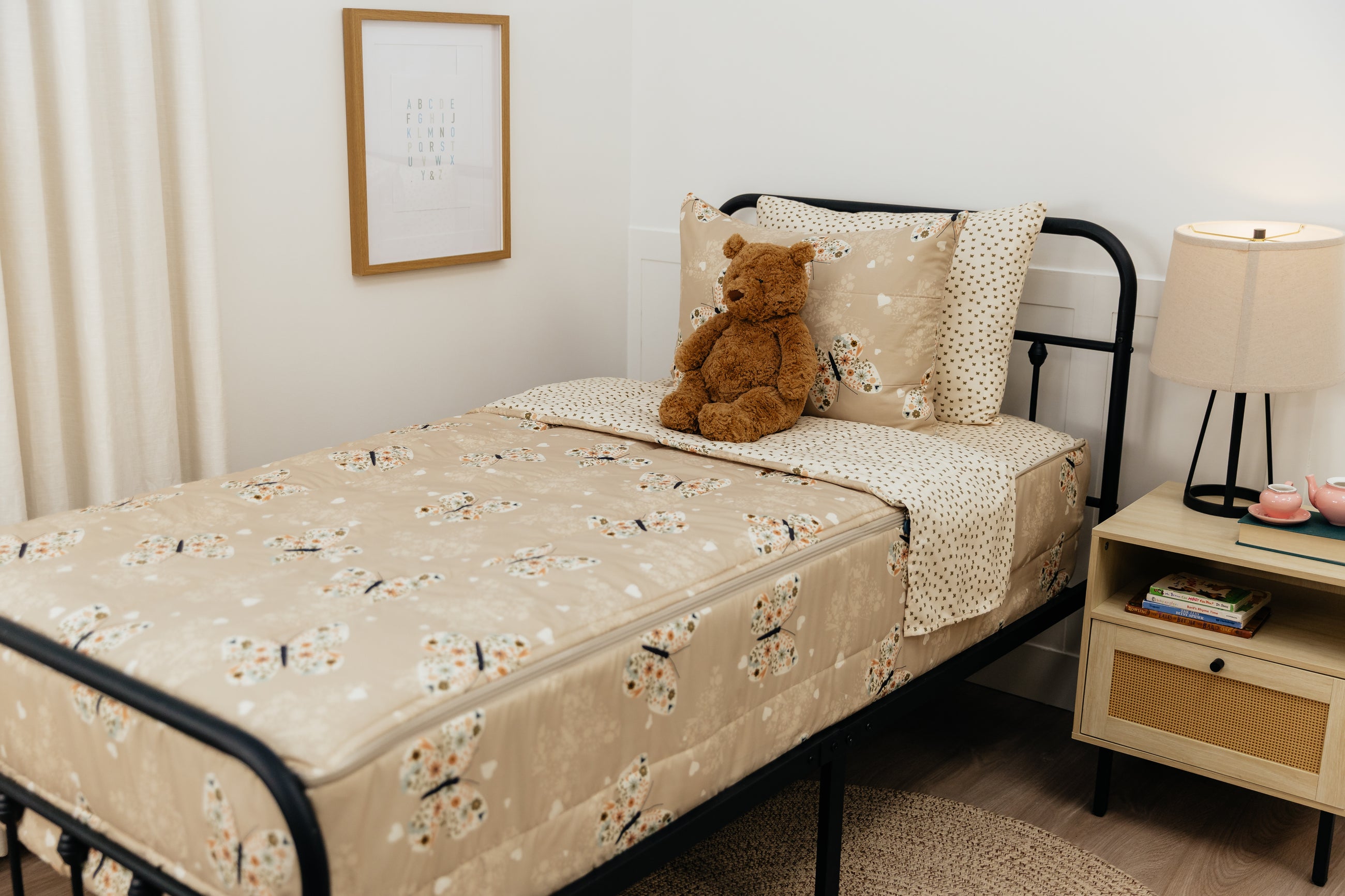 A twin bed made with butterfly zipper bedding, a nightstand beside the bed and a teddy bear propped up against the pillow.
