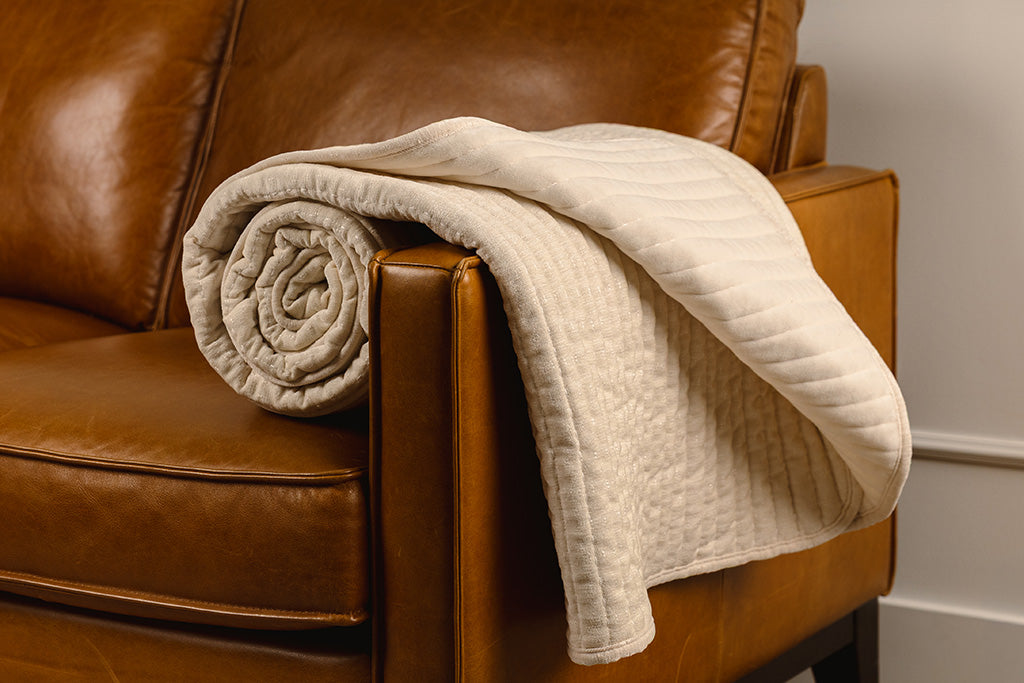 A cream chenille and minky blanket, rolled up on the arm of an orange leather couch.