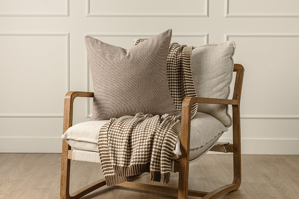A brown and cream scallop weave throw and a brown woven pillow on a wooden and oat colored chair.