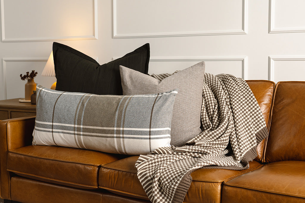 A black knit xl pillow, brown woven pillow, plaid xl lumbar pillow, and cream scallop weave throw on an orange leather couch.