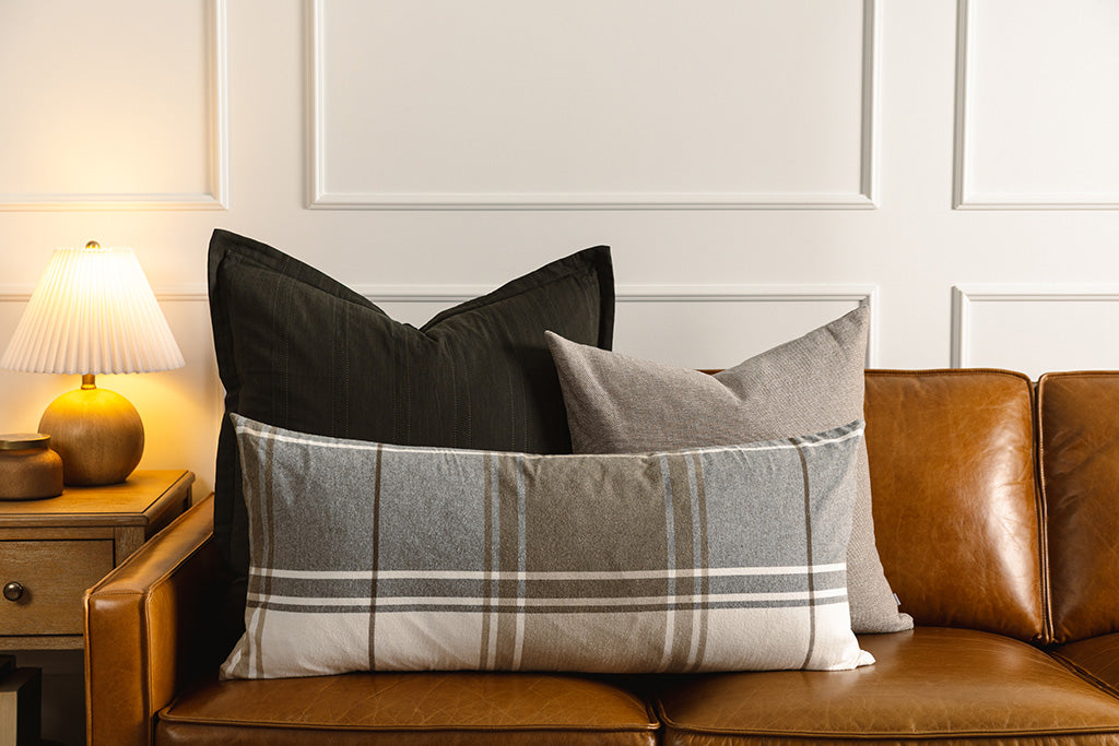 A black knitted pillow, brown woven pillow, and plaid lumbar on a leather couch.