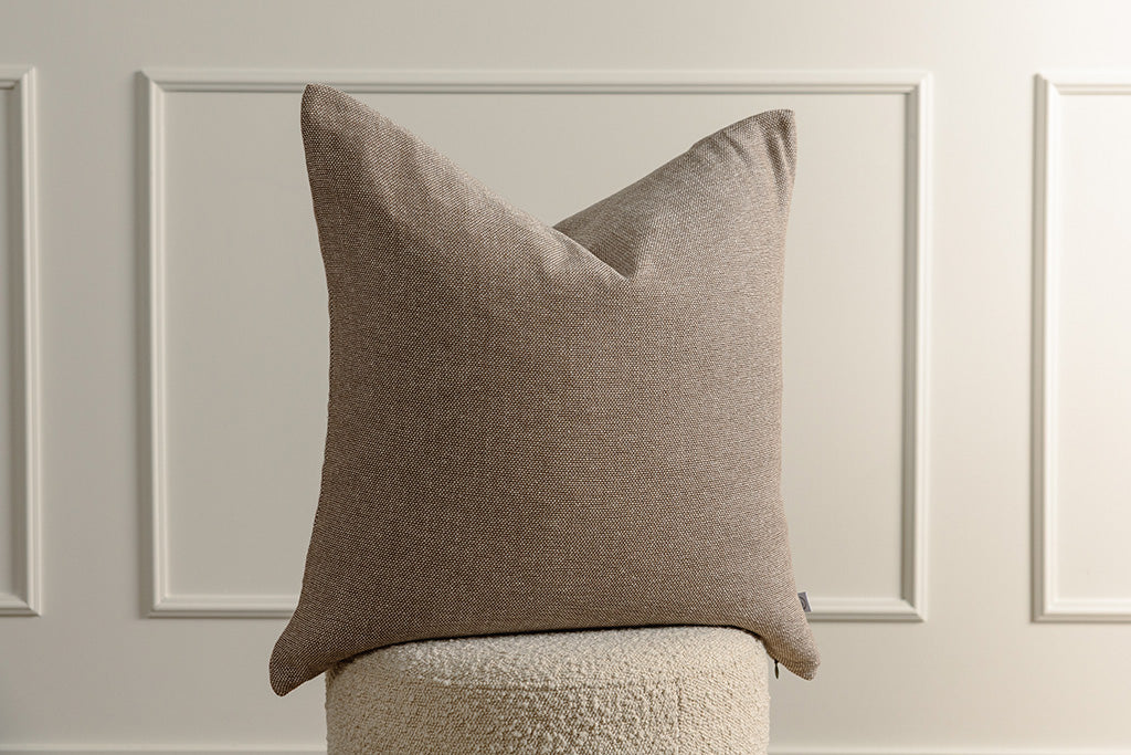 A brown woven pillow on a sherpa stool with a wainscoting background.