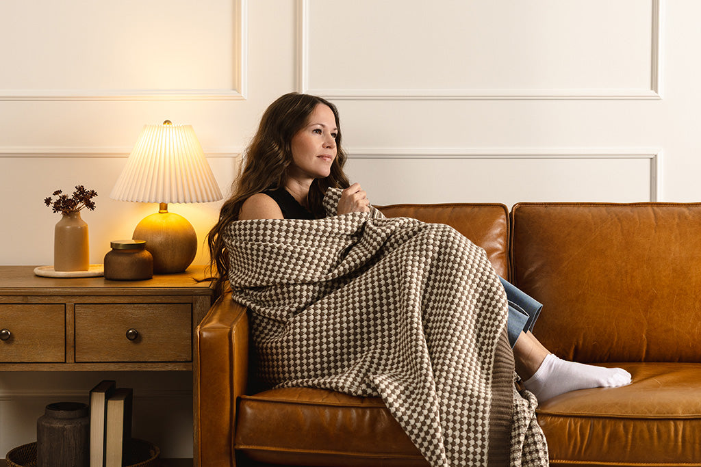 Woman wrapped in a brown and cream scallop weave throw on an orange leather couch.