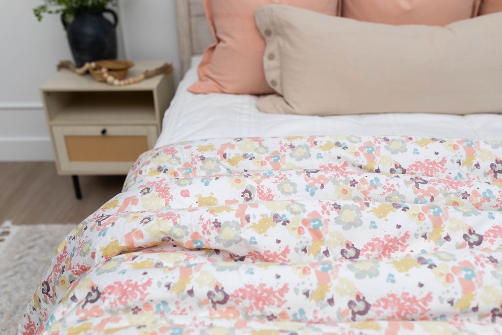 A close up of a bed made with white zipper bedding and neutral and peach pillows and a colorful floral pattern blanket.