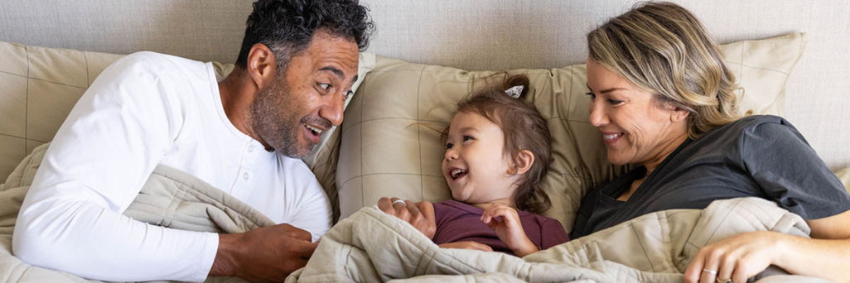Two parents and their toddler age daughter tucked into bed, laughing and smiling with one another.