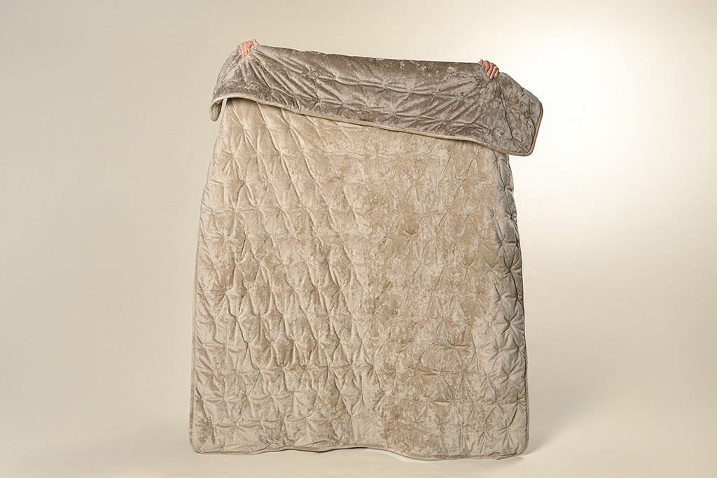A model holding up a tan and velvet textured blanket on a cream backdrop.