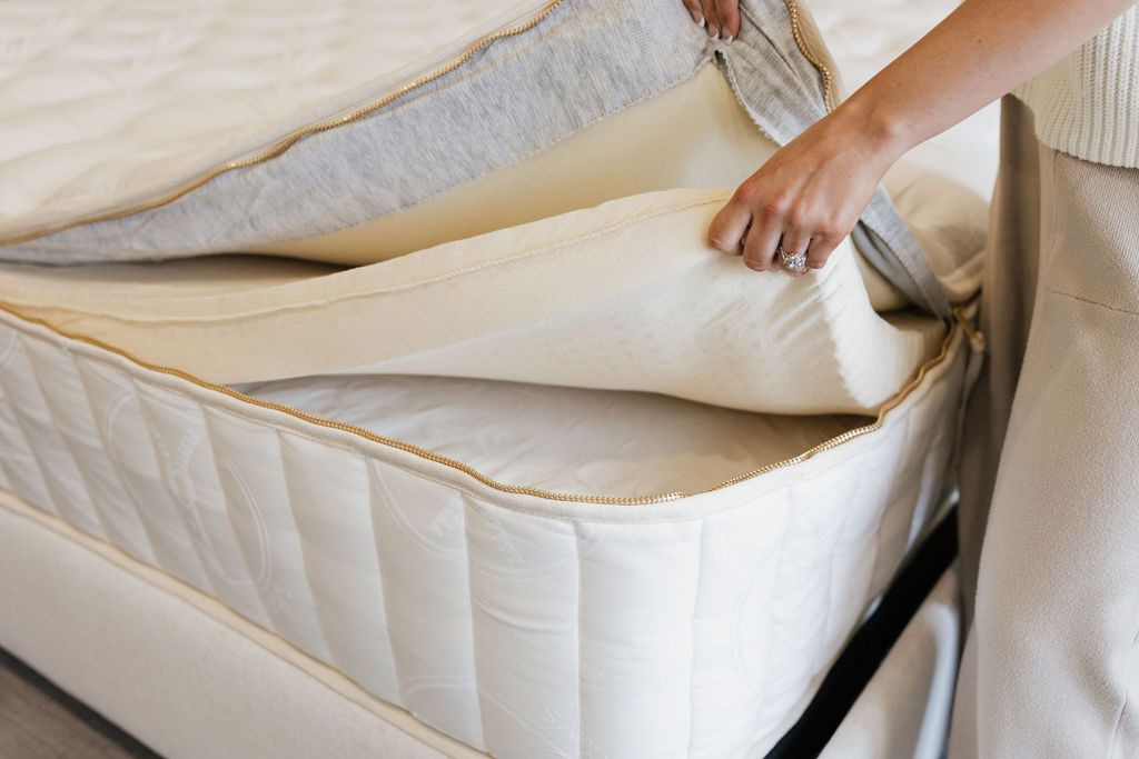 The inside of a zipper hybrid mattress, with a woman in tan pajamas lifting the comfort latex layer to reveal the coil layer beneath it.