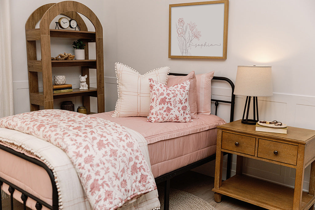 A bed made with pink zipper bedding and an assortment of pink floral and white tassel pillows and blanket.