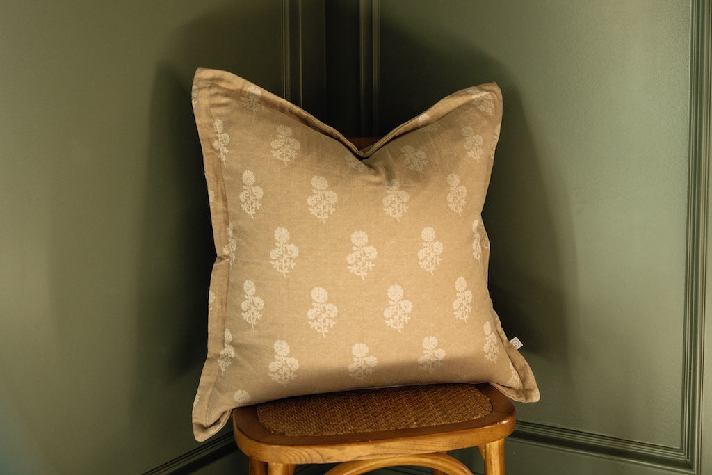 A tan and white floral pillow on a chair next to a green backdrop.