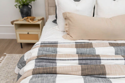 A close up of a made bed with white zipper bedding and an assortment of neutral pillows and a cream, gray, and tan plaid pattern blanket at the end of the bed.