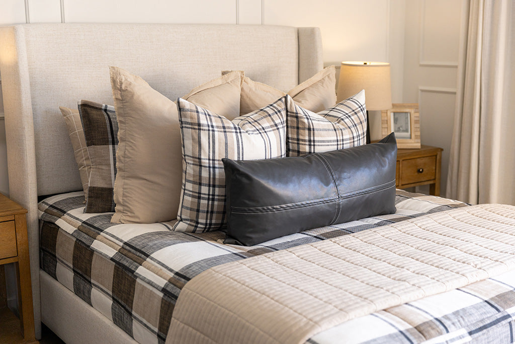 A close up view of a bed made with tan, cream, and charcoal zipper bedding and an assortment of neutral pillows and blanket.
