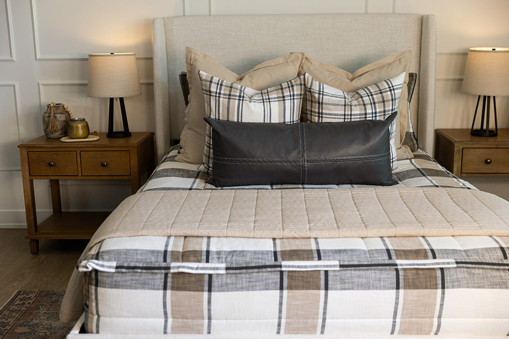 A bed made with cream, charcoal, and tan plaid zipper bedding and an assortment of neutral and leather throw pillows and blankets.