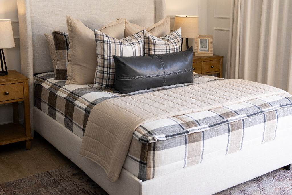 A bed made with cream, charcoal, and tan plaid zipper bedding and an assortment of neutral and leather throw pillows and blankets.