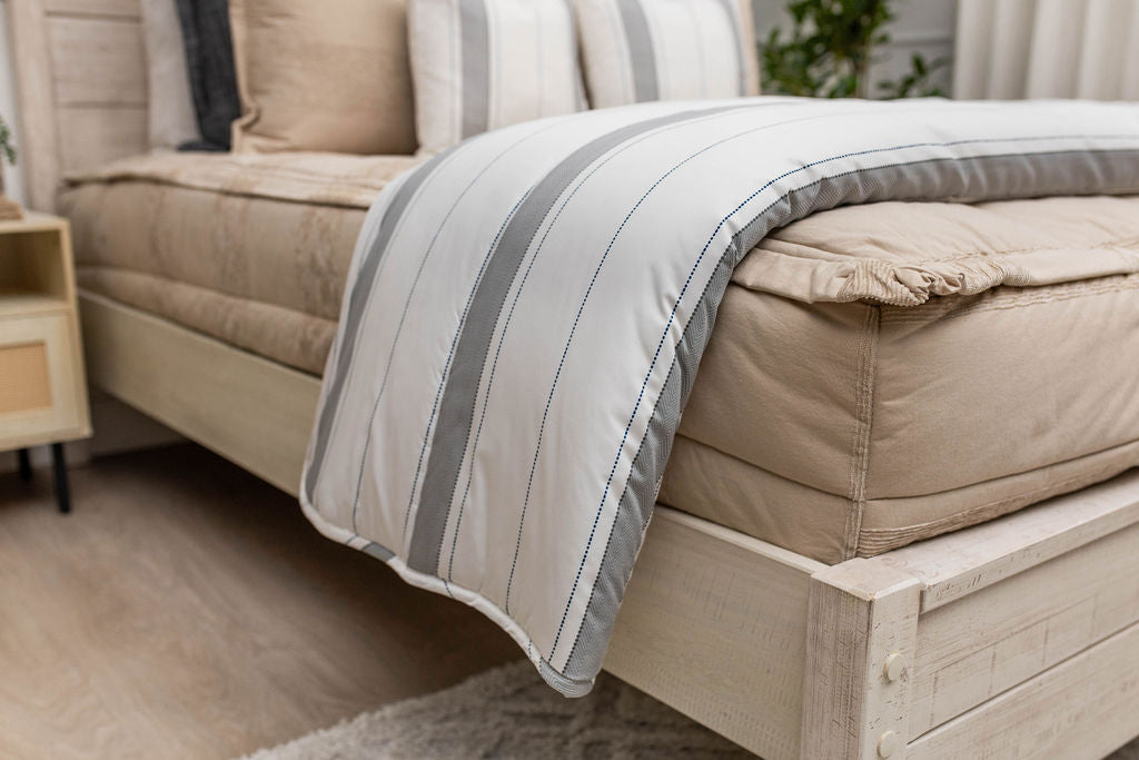 A side view of a bed made with tan zipper bedding with a white and gray striped blanket folded over the end of the bed.