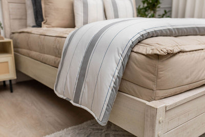 A side view of a bed made with tan zipper bedding with a white and gray striped blanket folded over the end of the bed.