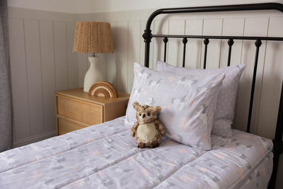 A twin bed made with purple unicorn zipper bedding, a nightstand beside it with a lamp and rainbow stacking toys