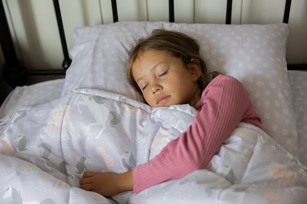 A young girl in pink pajamas sleeping in a bed made with purple unicorn kids zipper bedding