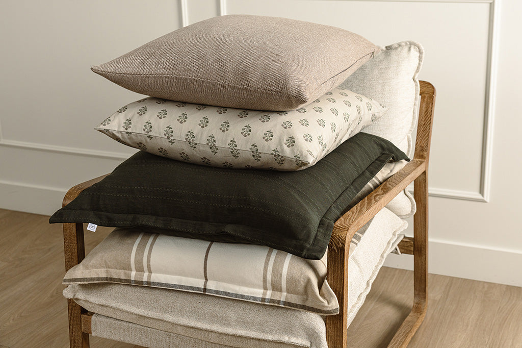 A plaid pillow, black knit pillow, floral blockprint pillow, and brown pillow stacked on a wooden and oat colored chair. 