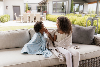 A brother and sister sitting beside each other and laughing together, both wrapped up in children's blankets.