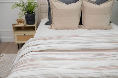 A close up of a made bed with cream embroidered zipper bedding and neutral pillows with a black and cognac striped white blanket at the end of the bed. 