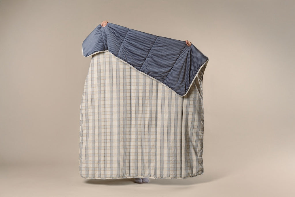 A blue and yellow plaid stitched blanket with a blue corduroy interior being held up by a model on a cream backdrop.