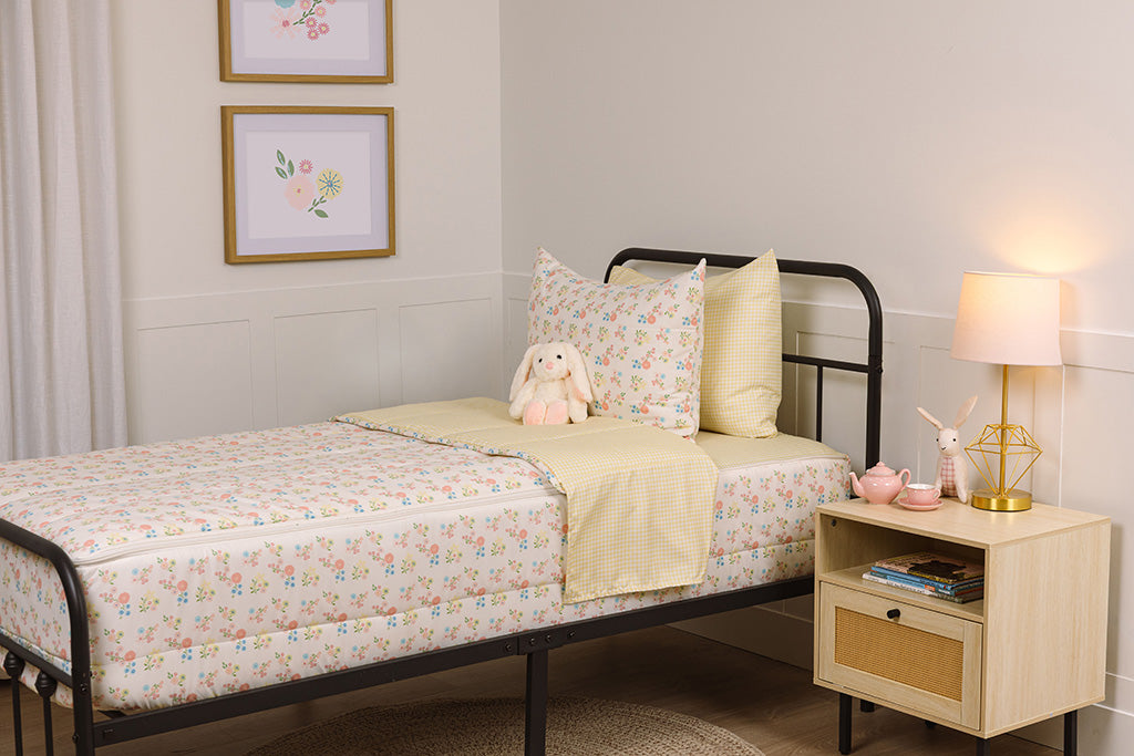 Children's zipper bedding in a bedroom on a black bed frame that has a flower print exterior and yellow gingham print interior, with a stuffed toy laying against the pillow.
