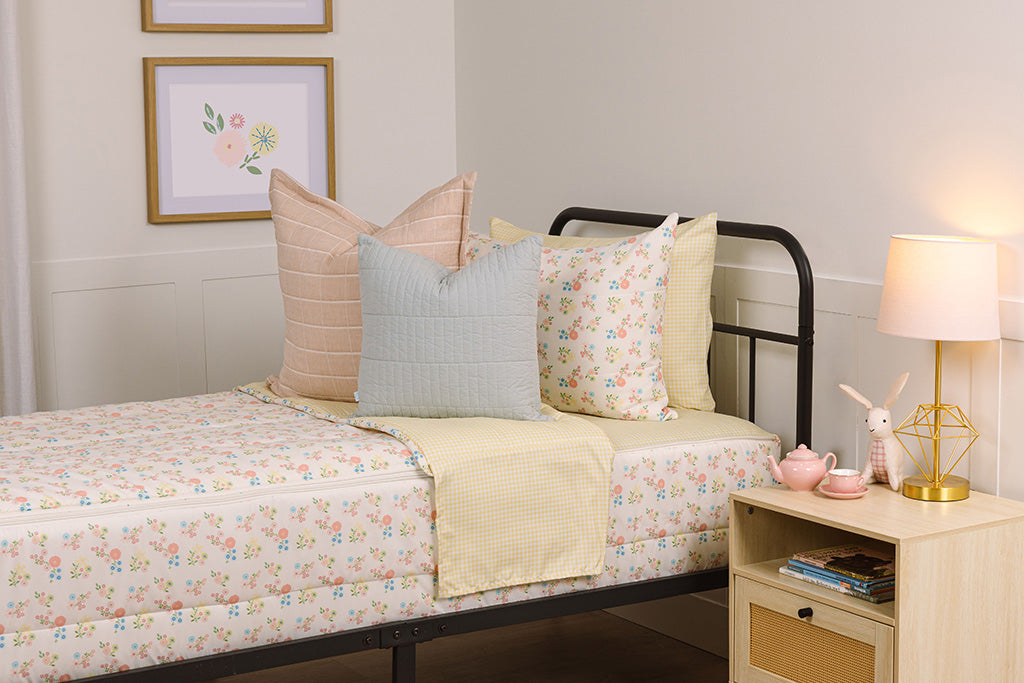 Children's zipper bedding in a bedroom on a black bed frame that has a flower print exterior and yellow gingham print interior, with pink and blue pillows.