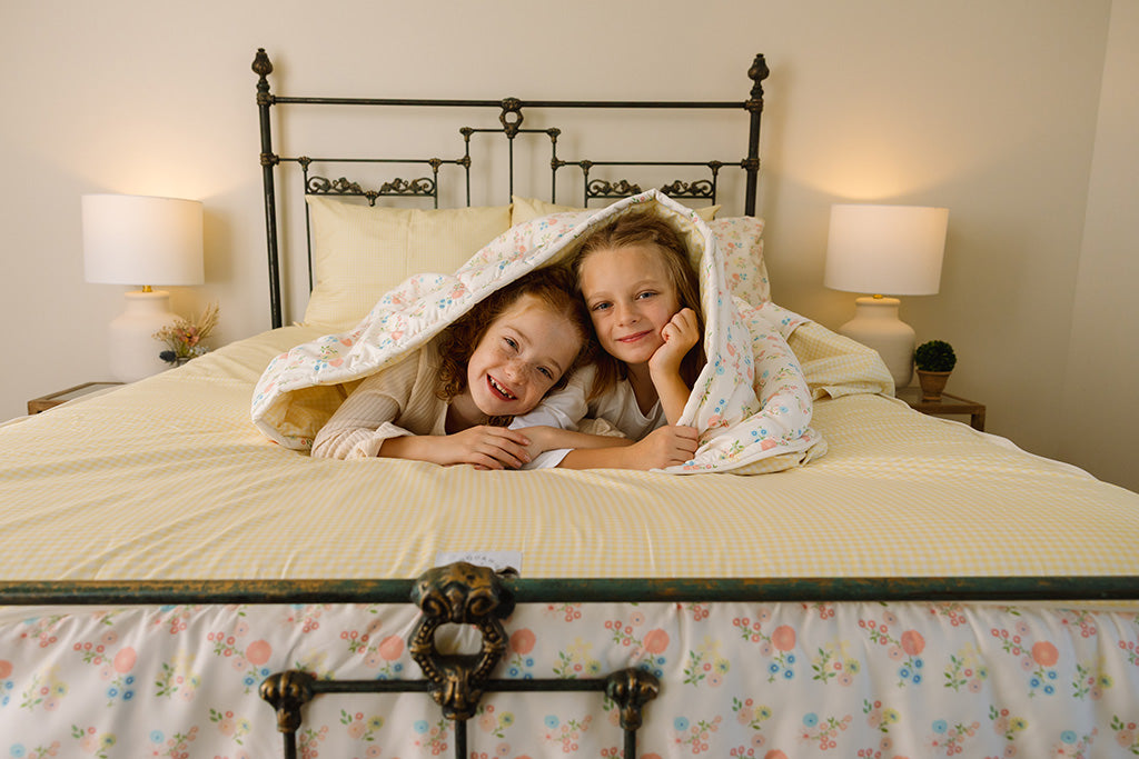 Two young girls lying next to each other under a floral duvet and yellow gingham zipper bedding.