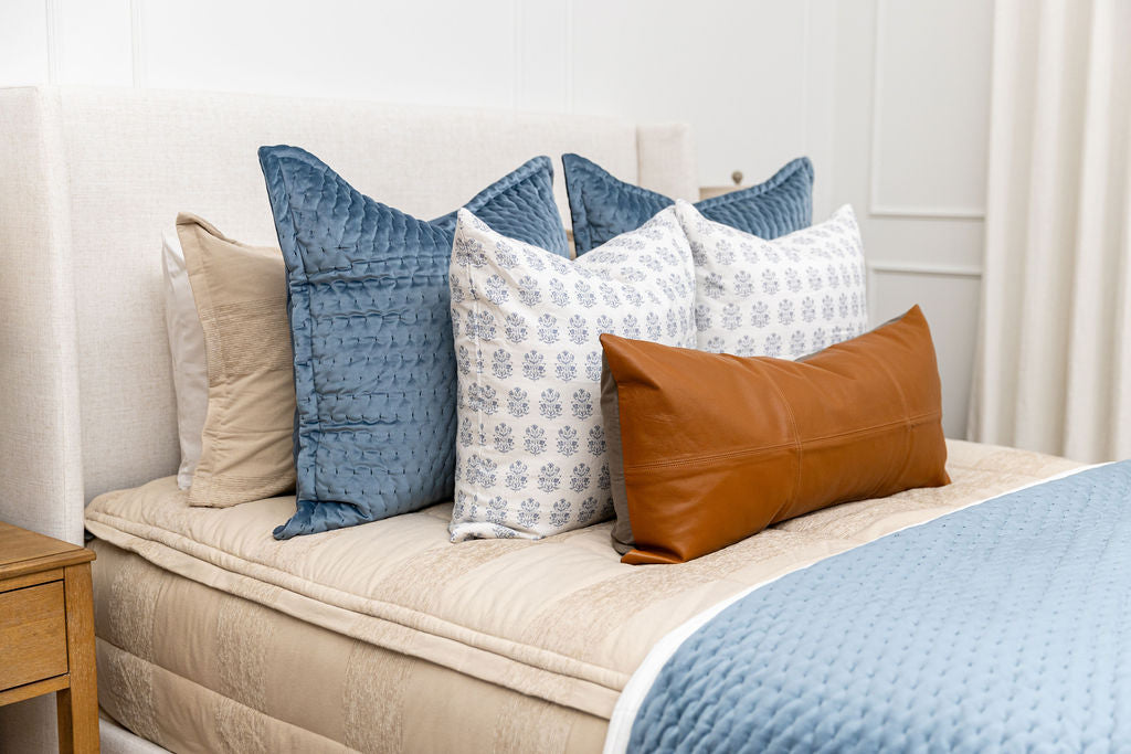 A queen bed made with tan zipper bedding and an assortment of blue and leather bedding accessories