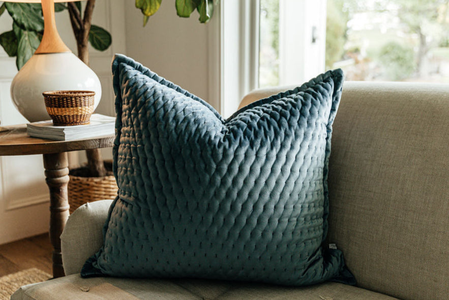 A blue velvet euro pillow arranged on a gray couch by a window and side table.