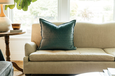 A blue velvet euro pillow arranged on a gray couch with a large window behind it and a side table.